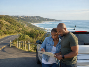 A South African couple standing beside their car at a quiet coastal lookout, checking a map or directions together before continuing their journey. The road stretches ahead in the background. The mood is calm and thoughtful — a natural picture of trusting guidance when you don’t know every step ahead.
