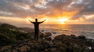 The silhouette of a person standing on the rocky South Coast shores at sunrise with arms wide open over the Indian Ocean, a gentle golden light radiating from their palms.